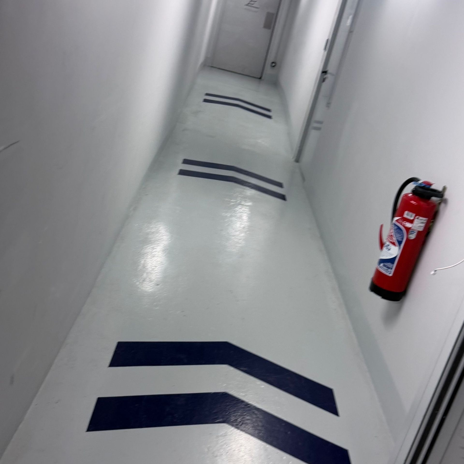 Couloir blanc étroit avec des flèches bleues au sol et un extincteur rouge fixé au mur