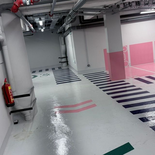 Parking souterrain avec sol blanc, bandes noires, canalisations au plafond et extincteur sur la gauche