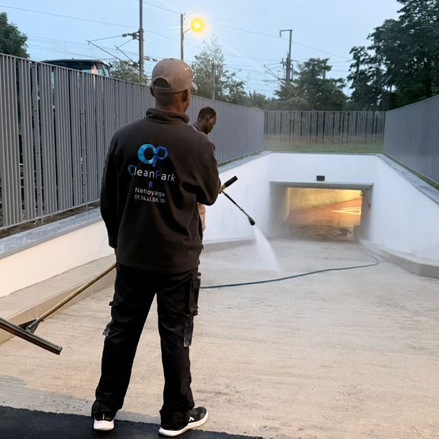 Deux ouvriers nettoient au nettoyeur haute pression une rampe de parking en pente près d'une entrée souterraine au crépuscule.
