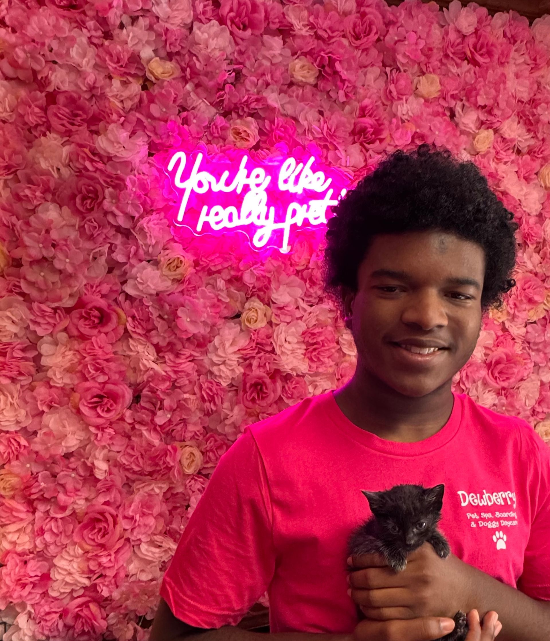 A man in a pink shirt is holding a black cat in front of a wall of pink flowers.