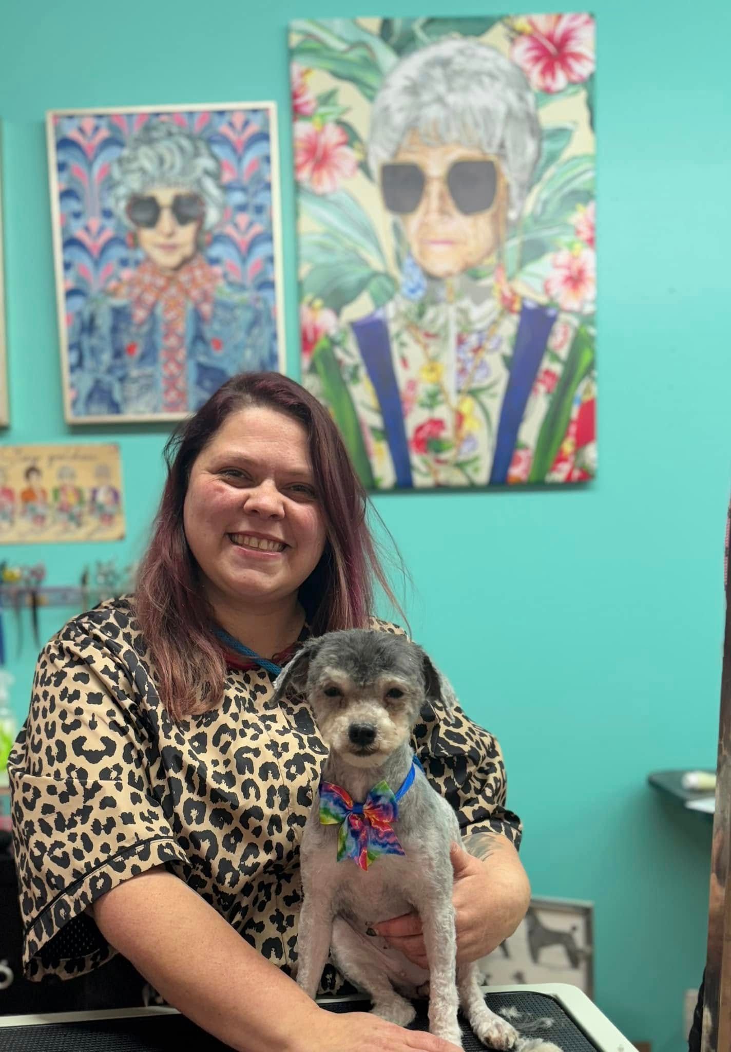 A woman is holding a small dog in front of a painting of an older woman.