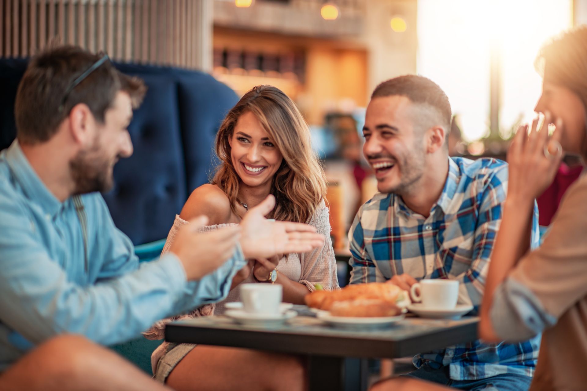 Four people laughing and talking around a table in a cafe, with food and coffee.
