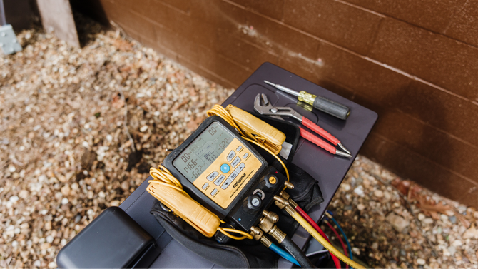 Electrician’s multimeter with probes and tools resting on a black panel beside a brown wall and gravel.
