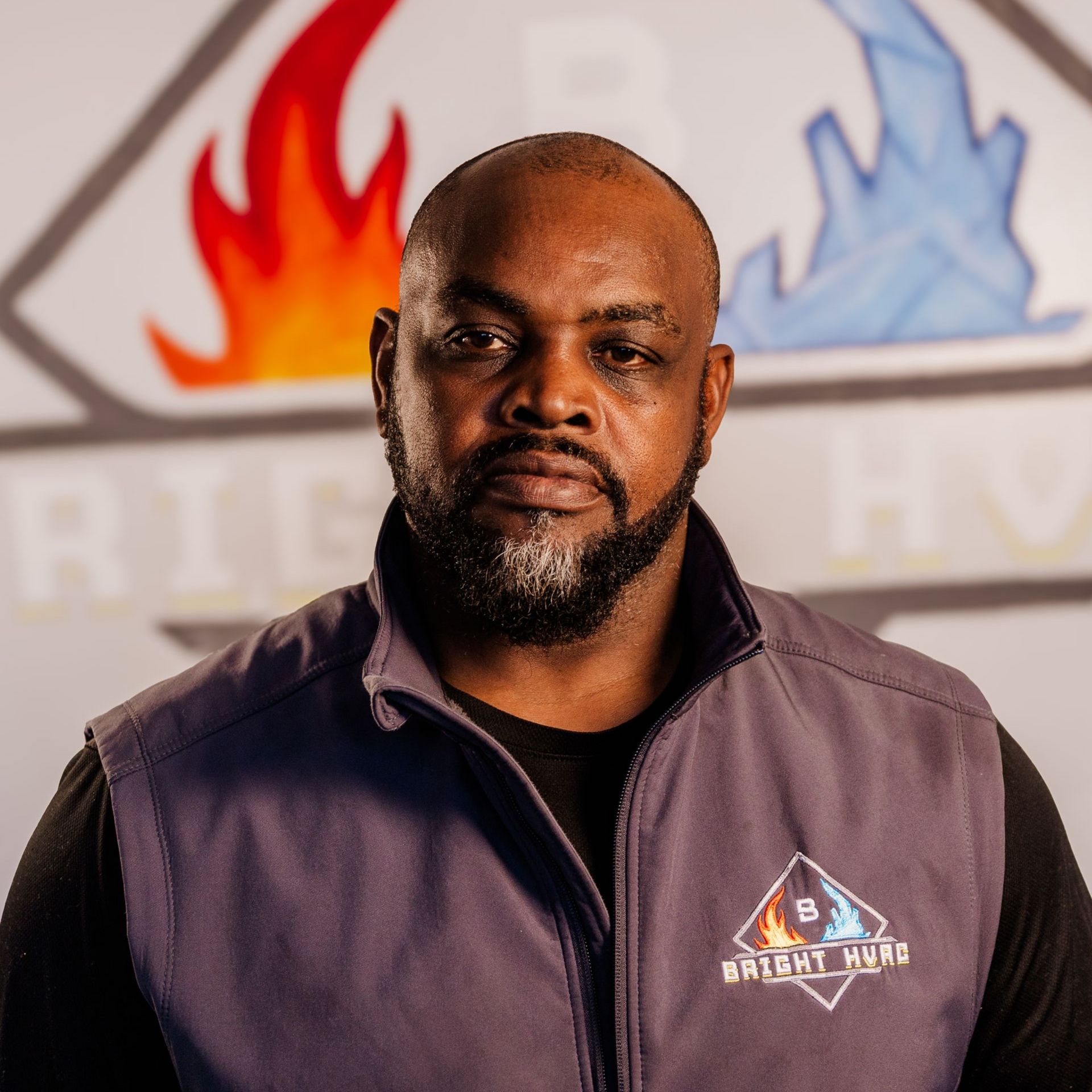 Portrait of a man in a purple vest with a logo, standing before a blurred flame and blue logo backdrop