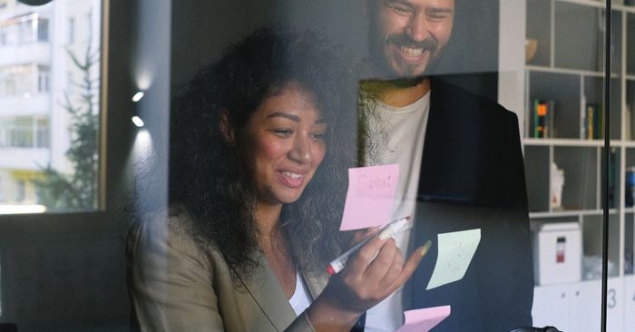 A man and a woman are looking at sticky notes on a glass wall.