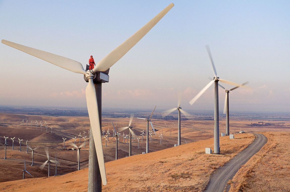 Turbine eoliche su una collina che generano elettricità. Paesaggio soleggiato e marrone con una strada.