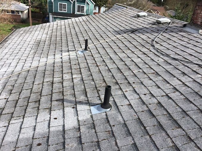 View of a gray shingled roof with three black pipes, a house in the background.