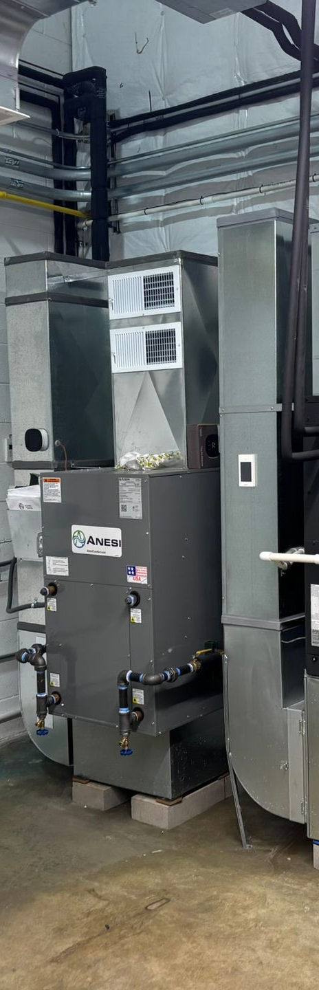 An industrial HVAC unit in a utility room. Gray metal units with various ductwork and pipes.