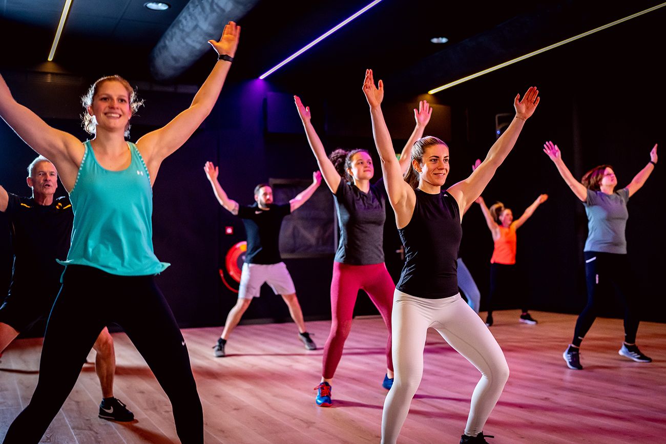 Een groep mensen met opgeheven armen tijdens een dansfitnessles in een studio met kleurrijke verlichting.