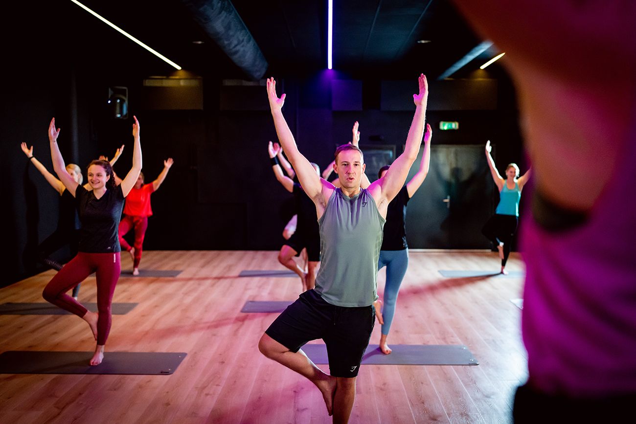 Mensen in een yogales, balancerend op één been met opgeheven armen, in een studio met gedempt licht.