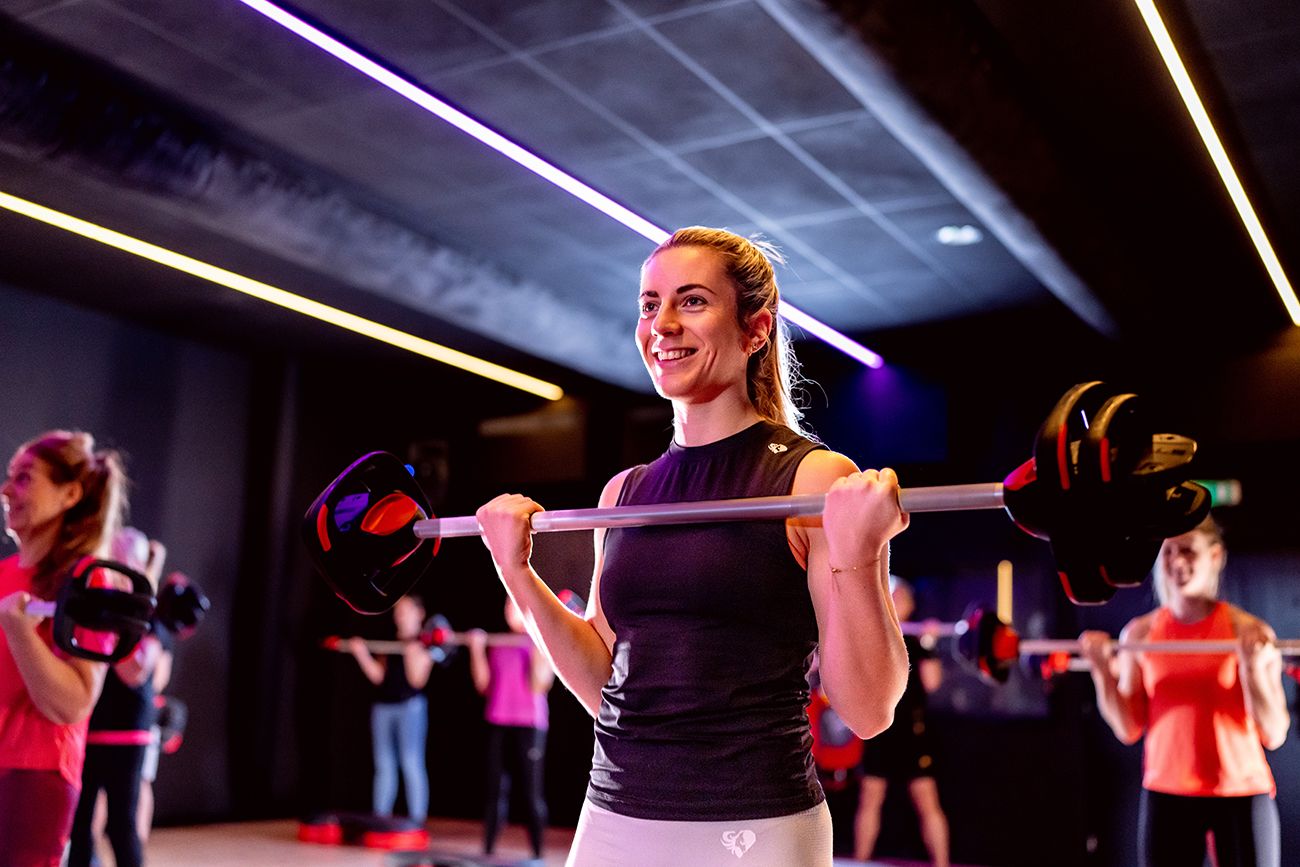 Een vrouw lacht terwijl ze een halter tilt tijdens een groepsfitnessles.