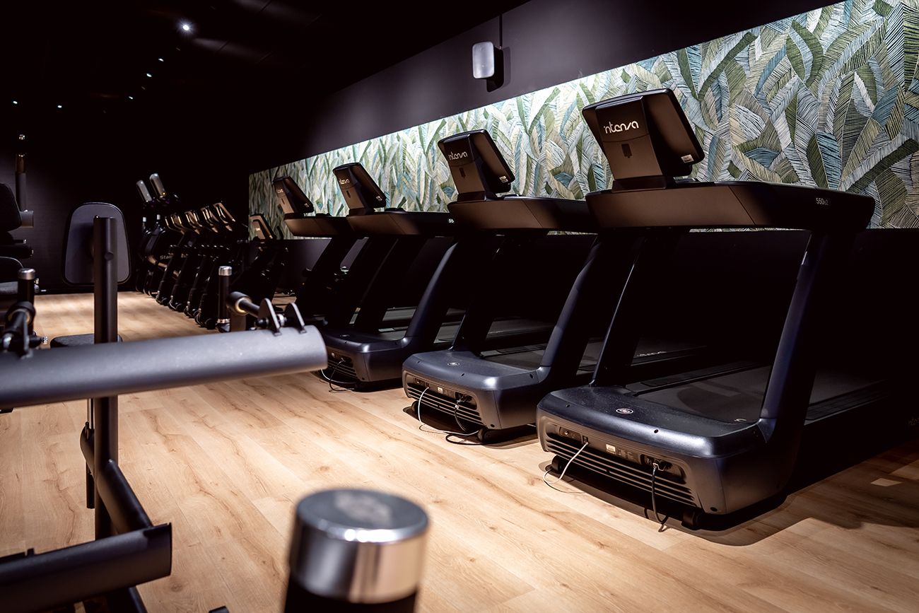 Rij loopbanden in een sportschool met een lichte houten vloer en een muur met patroon.