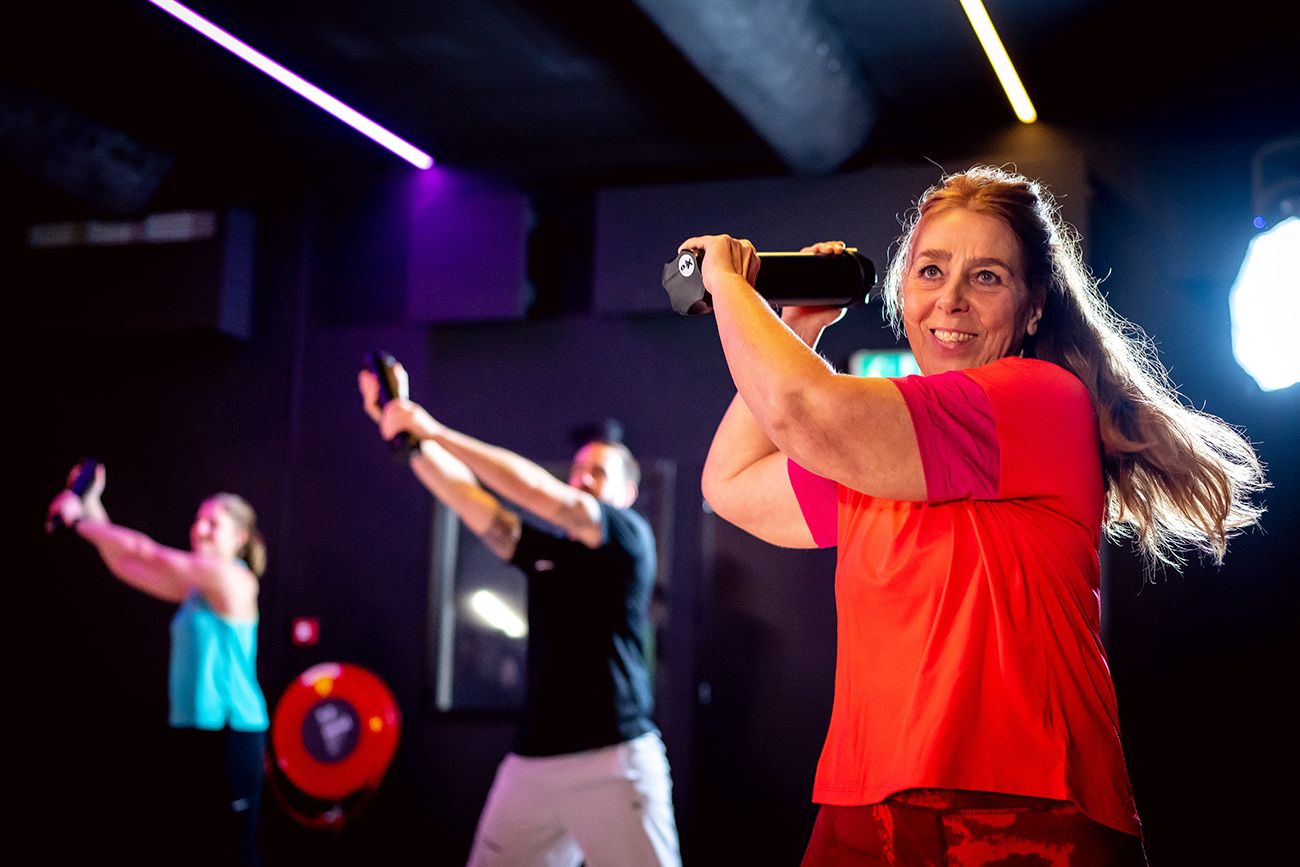 Mensen die binnen sporten met gewichten. Een vrouw in een oranje shirt lacht. Paarse en witte lichten.