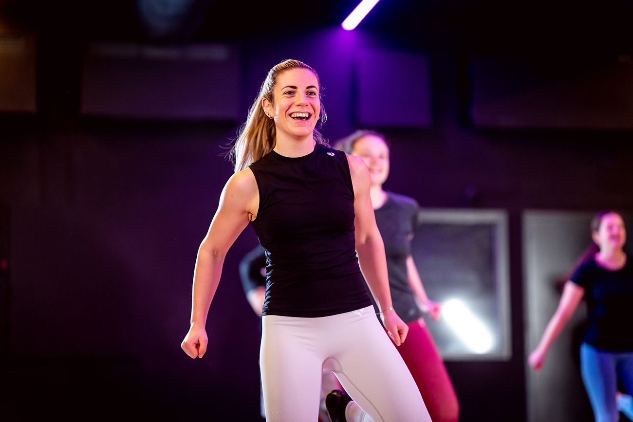 Een vrouw in sportkleding lacht terwijl ze samen met anderen in een studio aan het sporten is. Paarse verlichting.