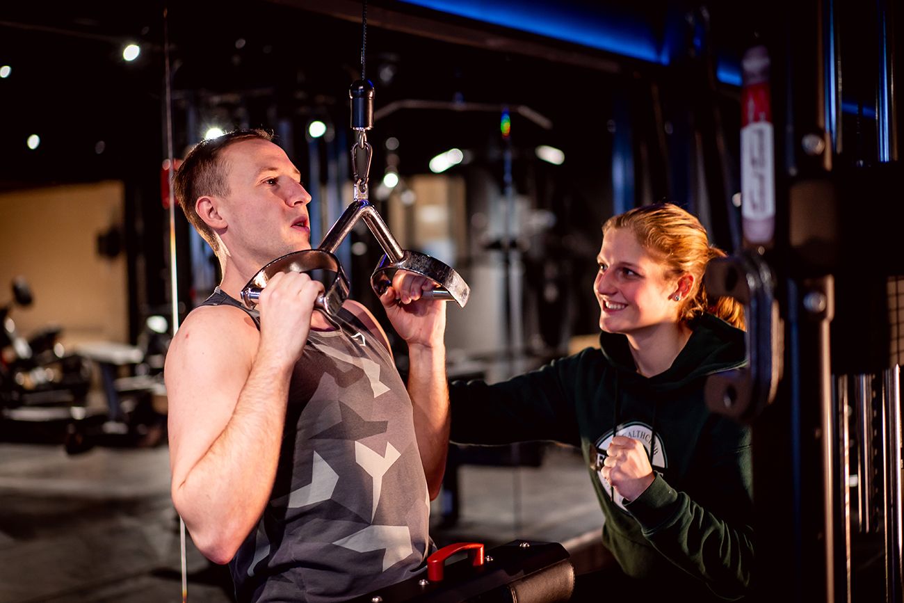 Een man en een vrouw in de sportschool. De man doet een lat pulldown, waarbij de vrouw hem assisteert.