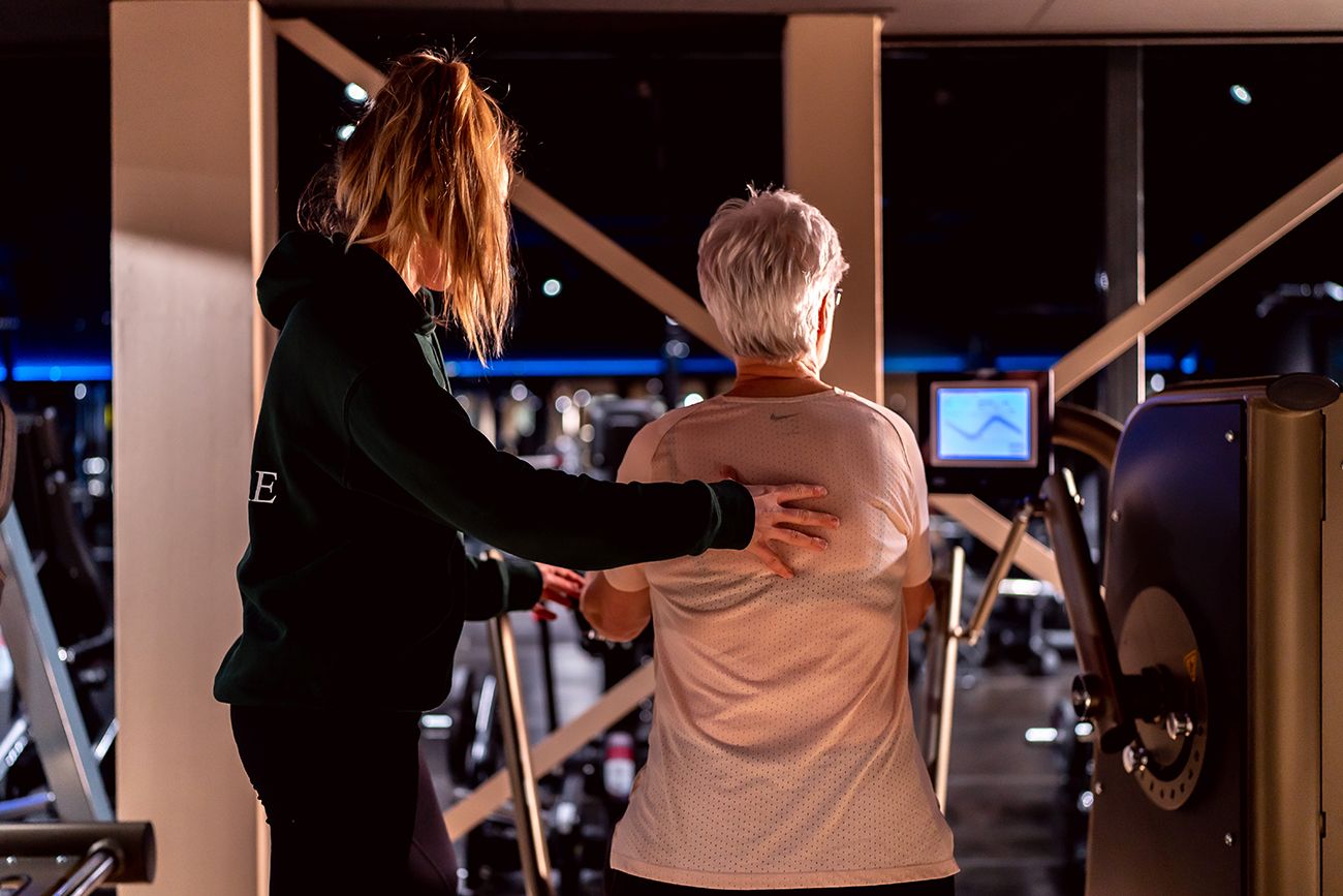Vrouw helpt oudere vrouw op crosstrainer in sportschool.