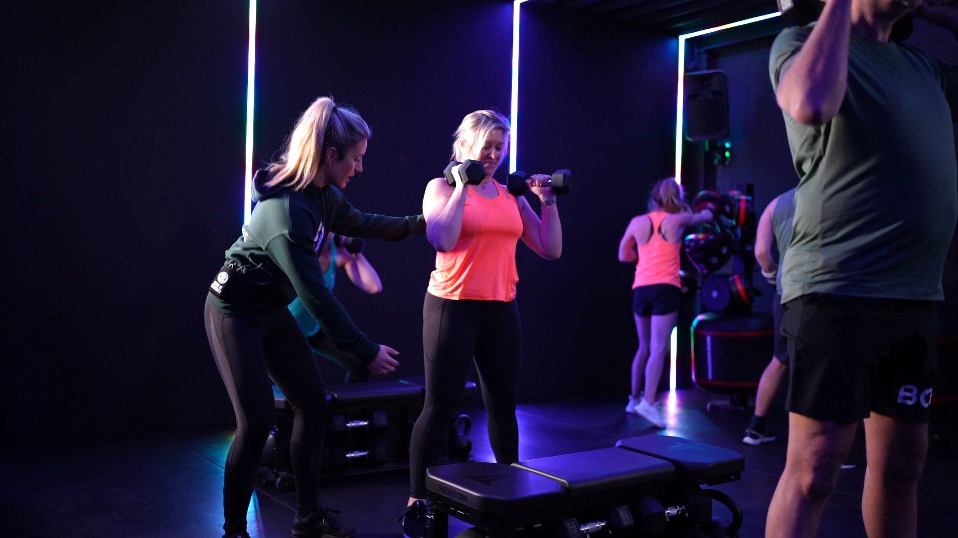 Mensen trainen in een sportschool met kleurrijke verlichting. Een vrouw in een oranje top doet squats