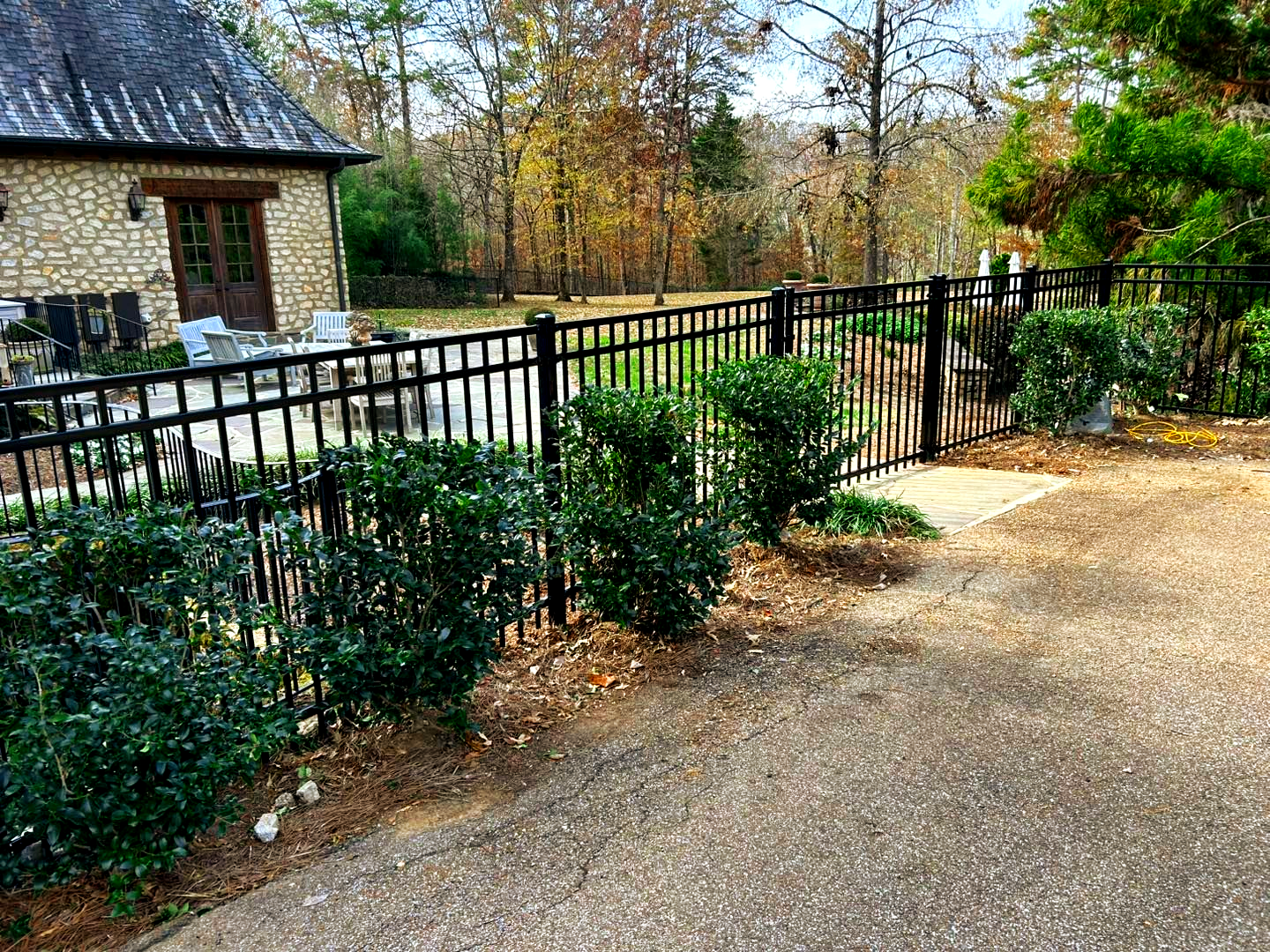 Black metal fence bordering a driveway, with bushes and a stone building in the background.