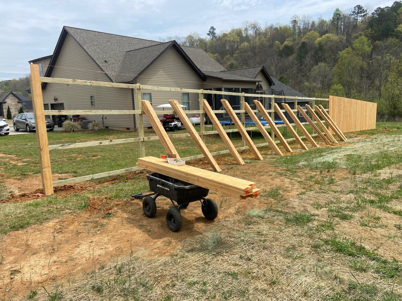 Fence construction in progress. Wooden fence posts and crossbeams being assembled in a yard with a house in the background.