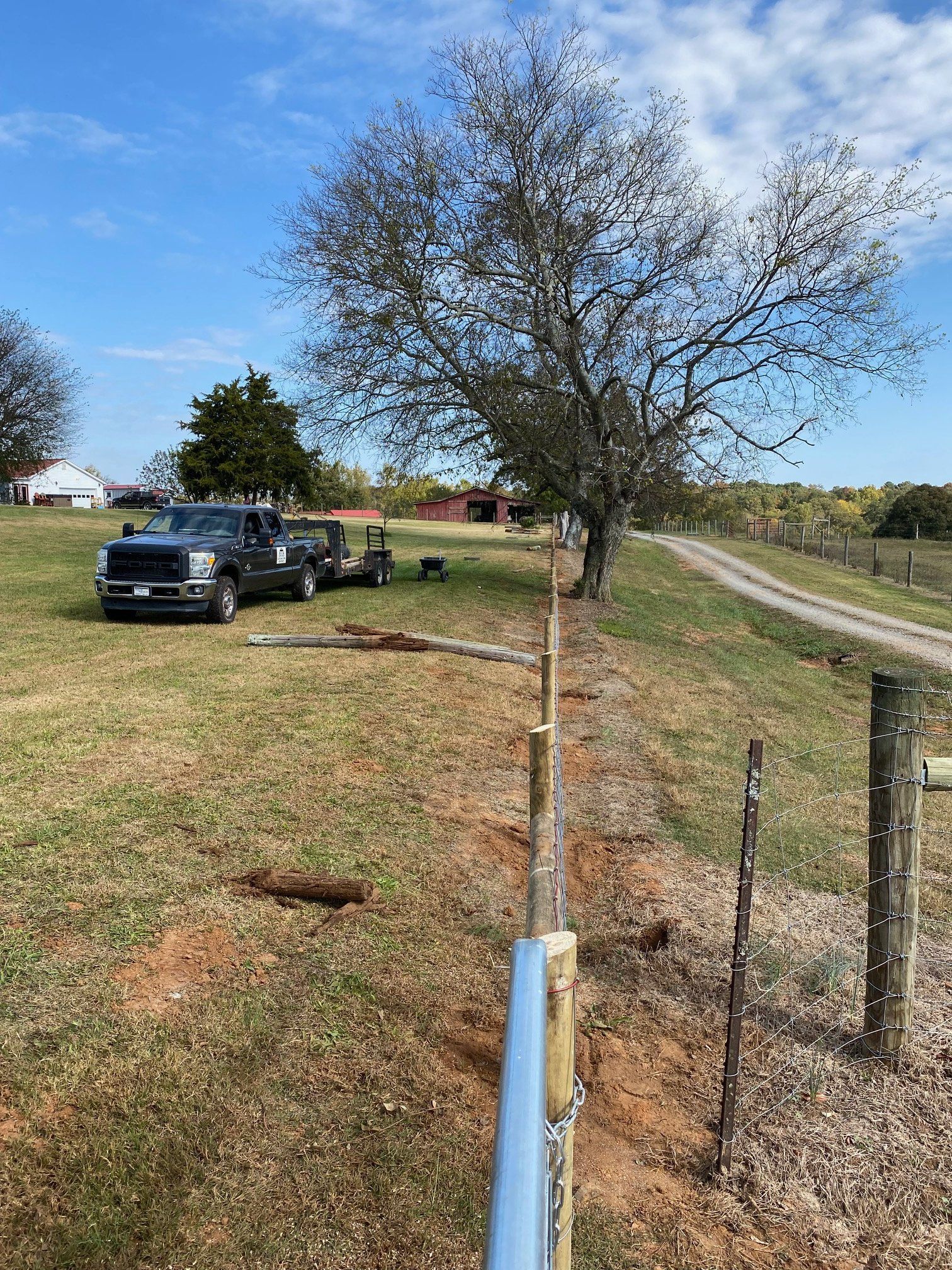 Fence being built in a rural setting with a truck, trailer, and tree under a blue sky.