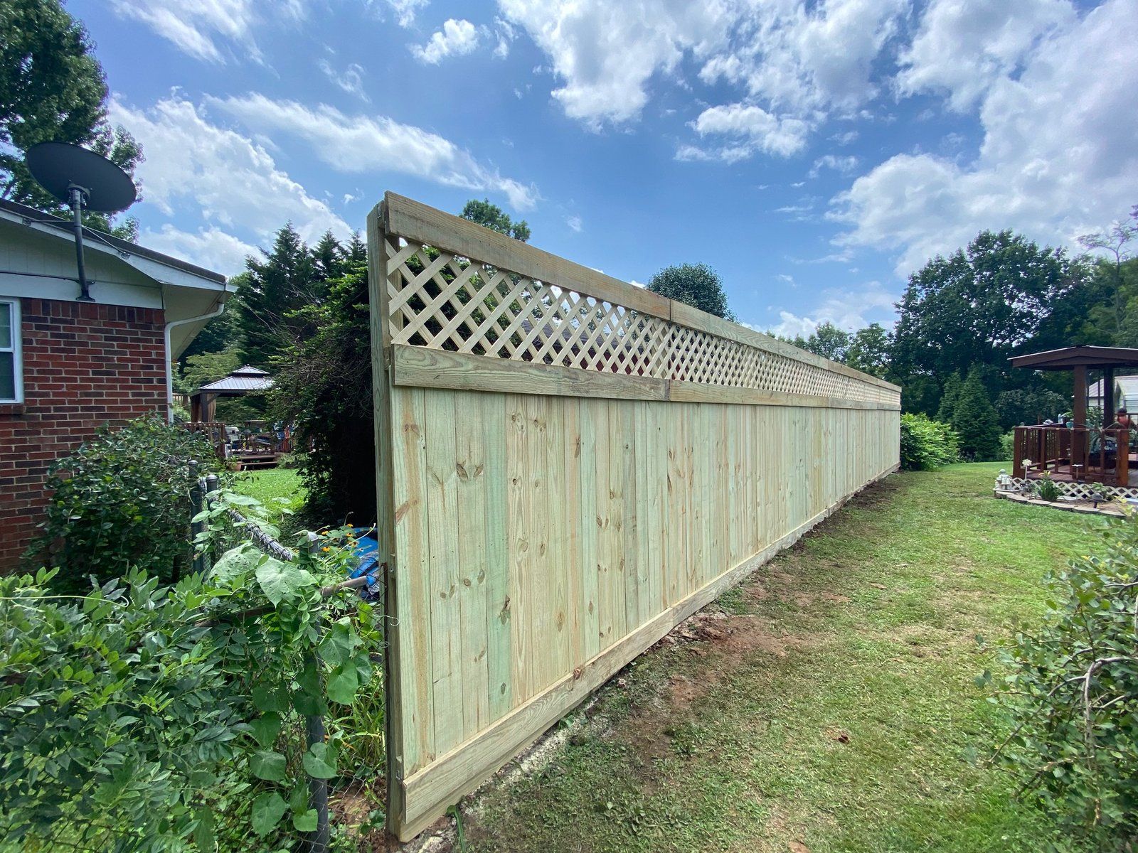 Wooden fence with lattice top in a backyard, under a blue sky with clouds.
