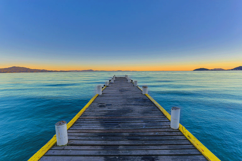 Cena de praia com prédios, oceano e montanhas sob um céu azul. Pessoas caminhando na areia.