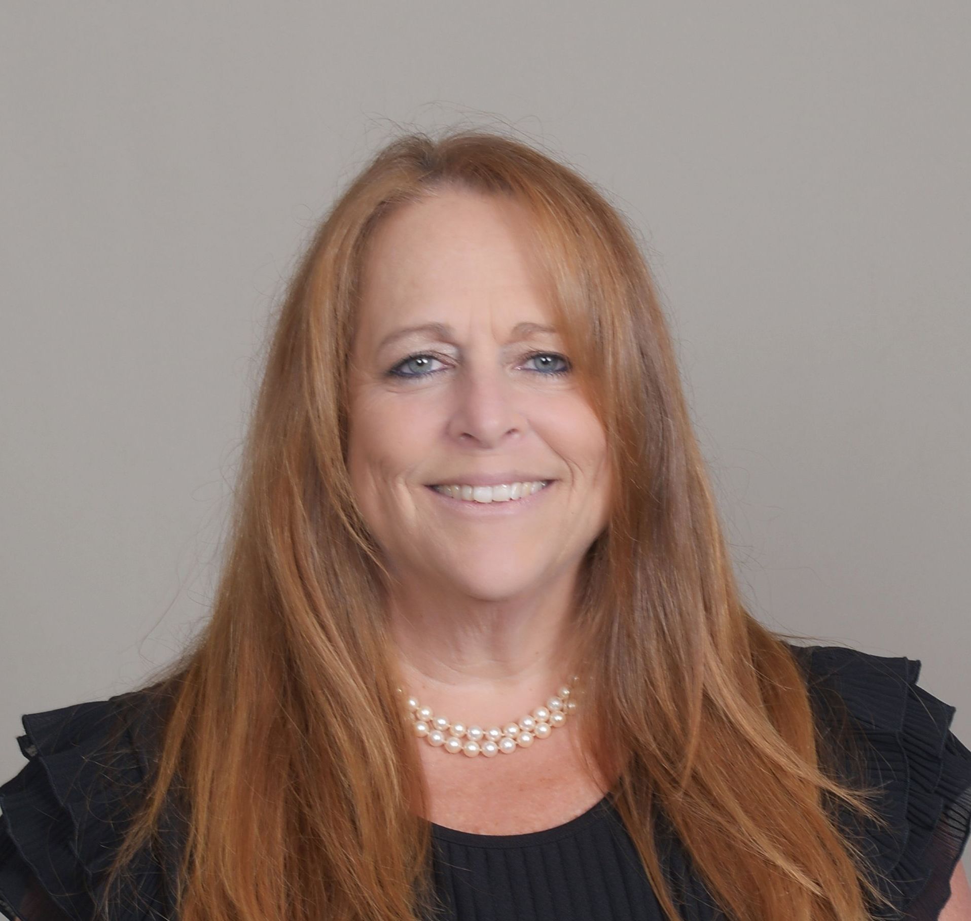 A woman with long red hair and a pearl necklace is smiling for the camera.