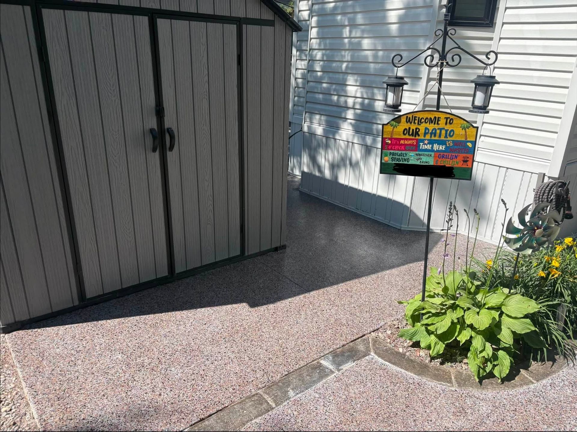 Small colorful sign beside a house and garage, next to a leafy plant in a sunny yard.