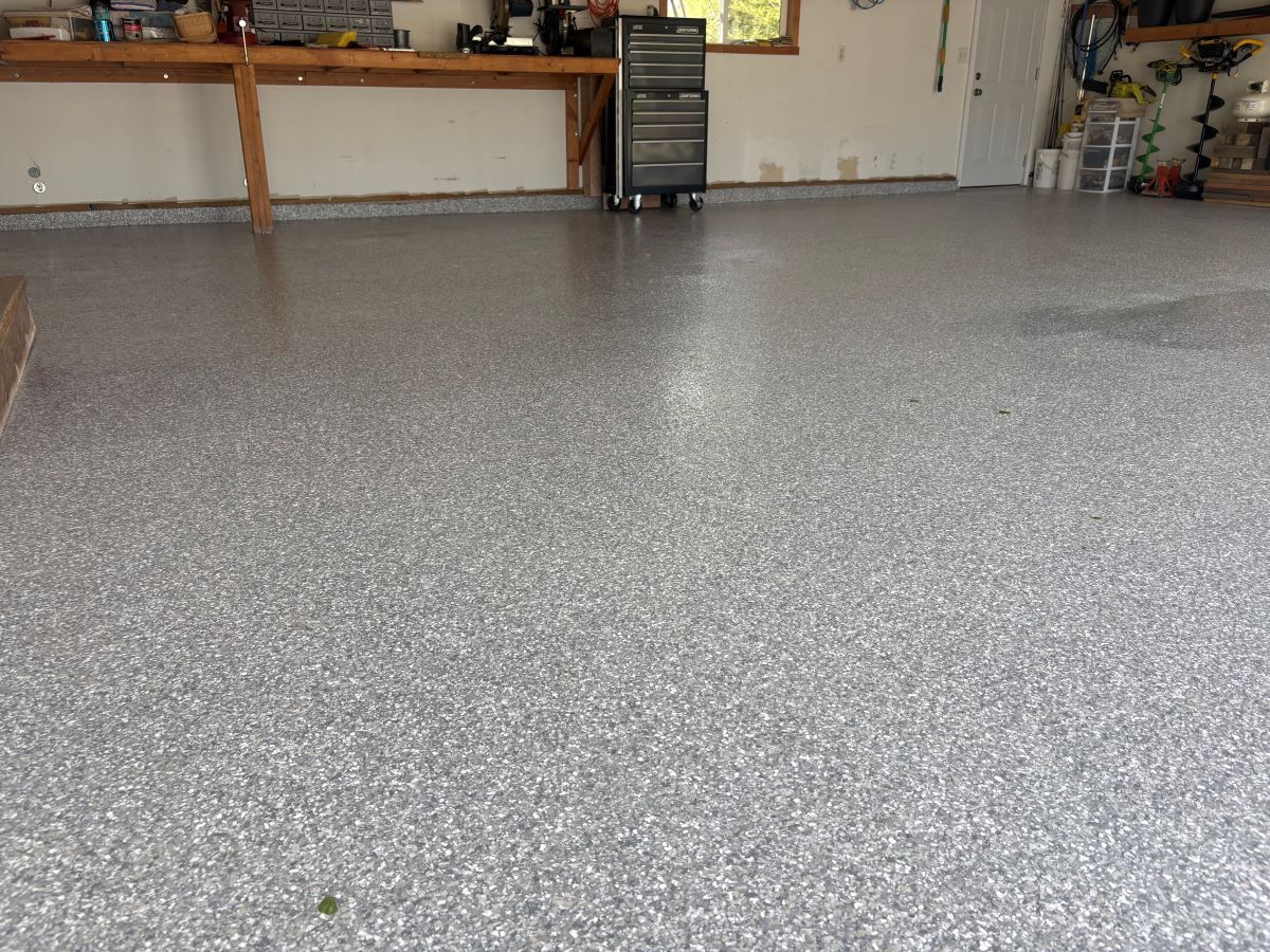 Empty speckled garage floor with wooden workbench and storage shelves along the back wall