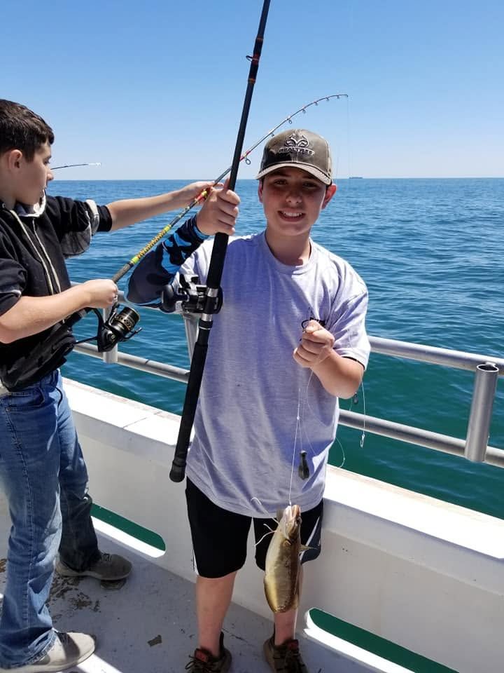 Person on a boat holds up a fish while another person reels in a fishing rod on calm blue water.