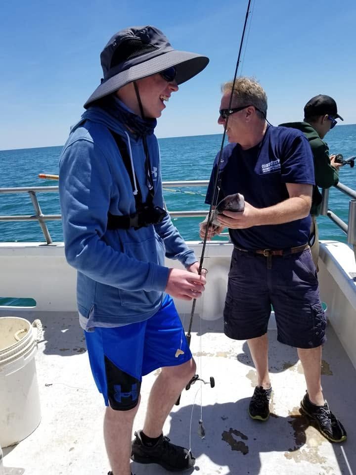 Two men on a boat holding fishing rods, one in a blue sun hat and shirt, with the ocean behind them