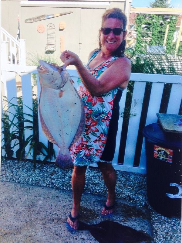 Woman in sunglasses holding a large fish on a dock, wearing a colorful dress and flip-flops.