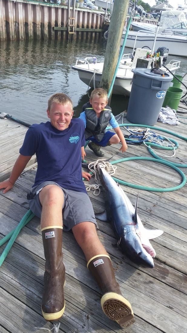 Two people sit on a dock beside a large fish, with coiled hoses and water in the background.