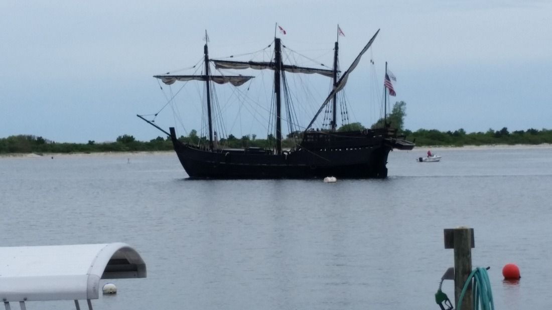 Black sailing ship on calm water near shore, with masts and flags under a cloudy sky.