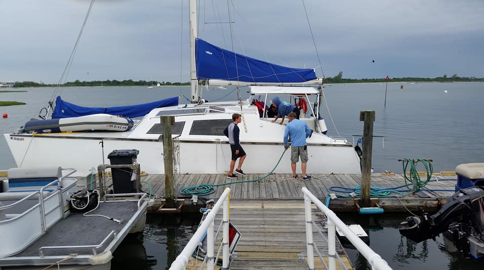 People boarding a white sailboat at a marina, with a blue sail cover and calm water in the background