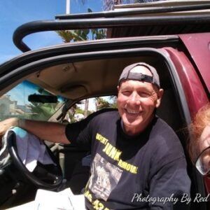 Man in black shirt smiles, sits in red truck. Gray cap, window in background.