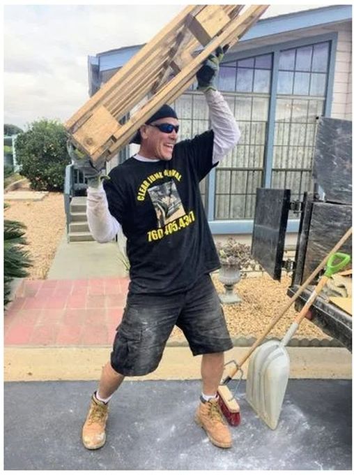 Man lifting a wooden pallet. Wearing black shirt, shorts, work boots, sunglasses, and a black hat. Outdoors.