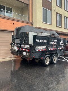 Black trailer loaded with junk, parked in front of an apartment building in the rain; phone number on trailer.