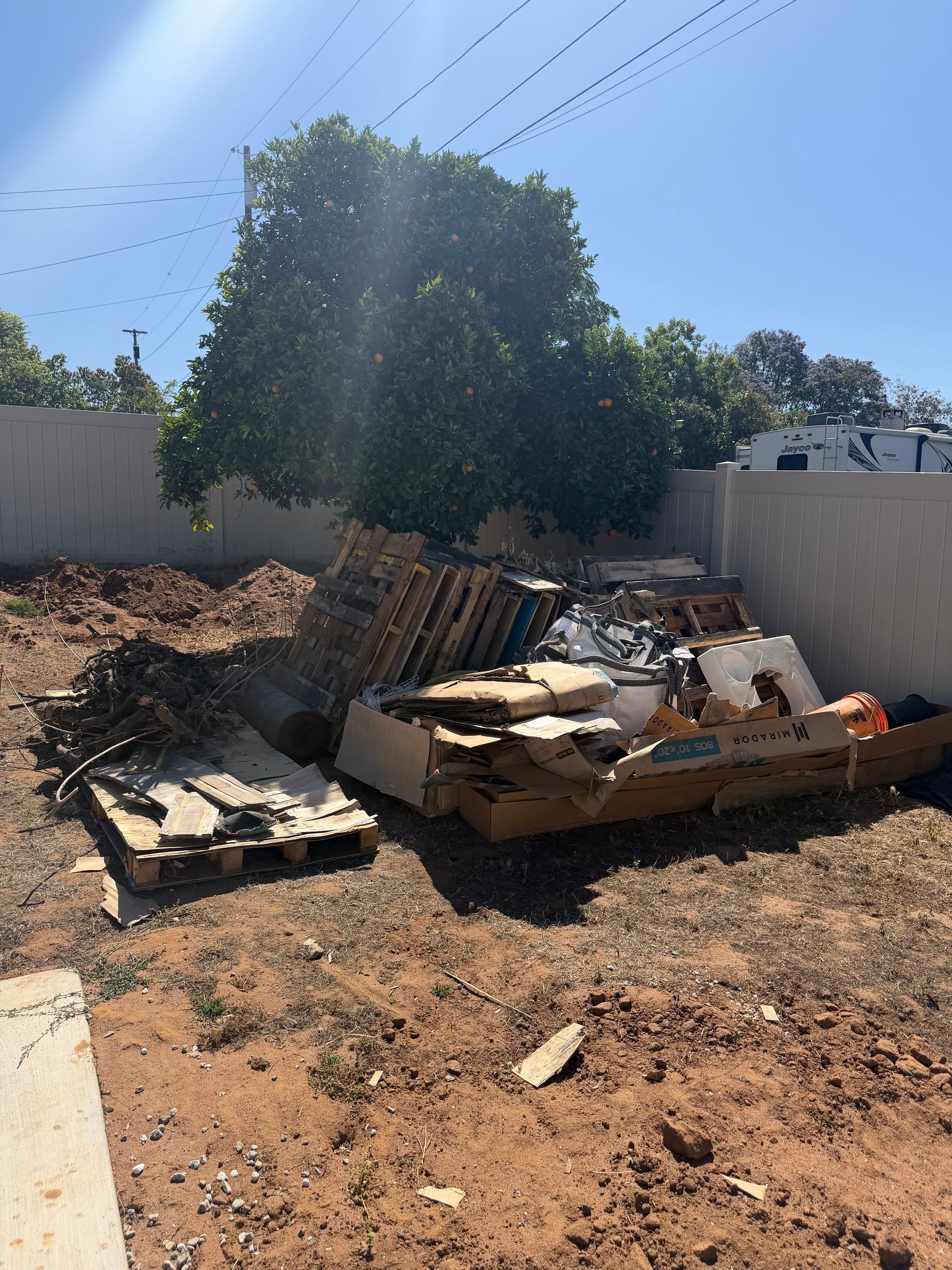 Piles of wood in a parking area with a building and trees in the background. A sign is visible.