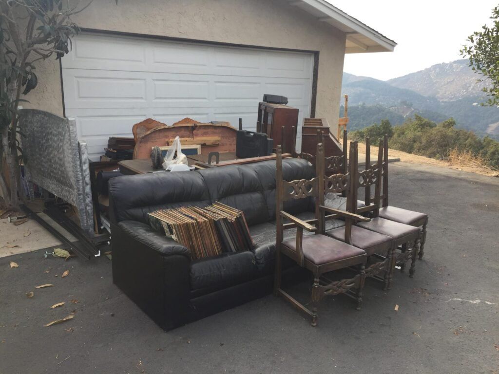 Furniture piled outdoors in front of a garage: sofa, chairs, cabinets, with a mountain view.