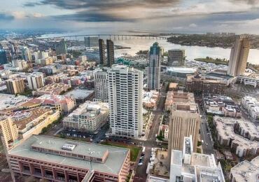 Aerial view of San Diego downtown cityscape with the Coronado Bridge visible in the background under a cloudy sky.