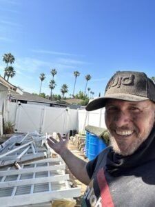 Man in hat smiles, gesturing toward pile of white fence parts with a house and palm trees in the background.