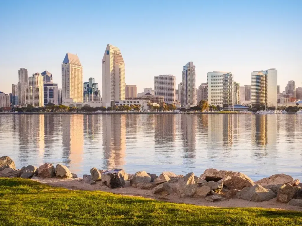 San Diego skyline reflected in calm water with a grassy bank and rocks in the foreground.