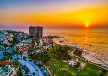 Sunset over a coastal city with a tall building, palm trees, and ocean.