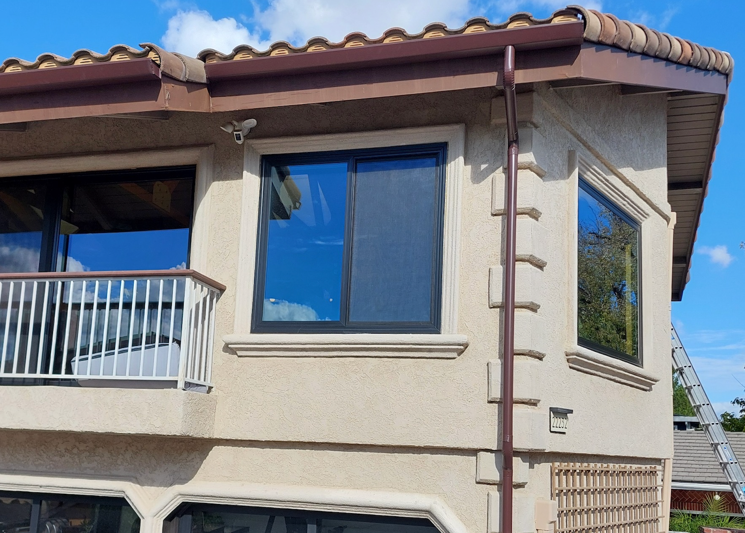 Corner of a beige stucco house with blue-tinted windows, white balcony railing, and a tiled roof under a clear sky
