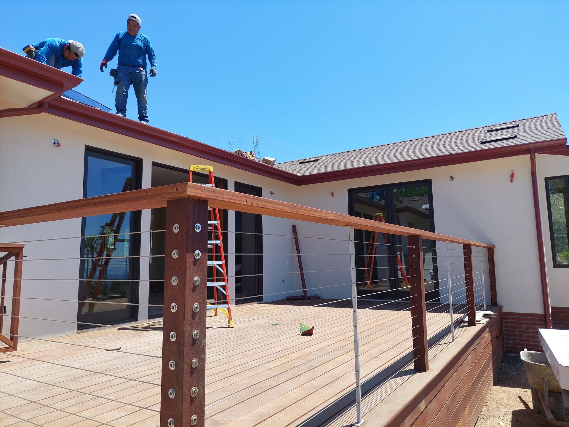 Worker on a rooftop beside a house under construction, with deck railings and a ladder below.