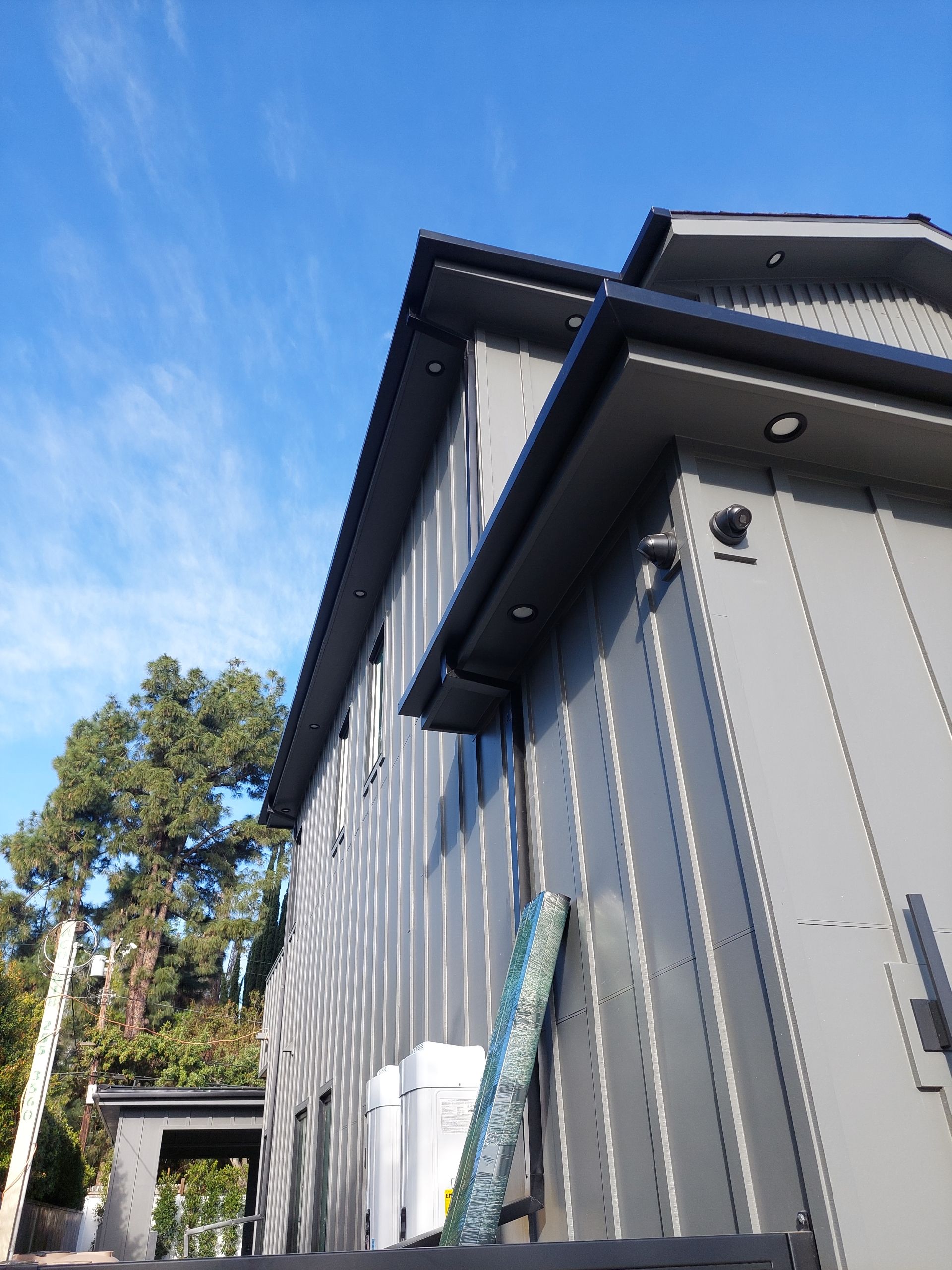Gray house exterior with tall siding, dark roof trim, and blue sky above trees