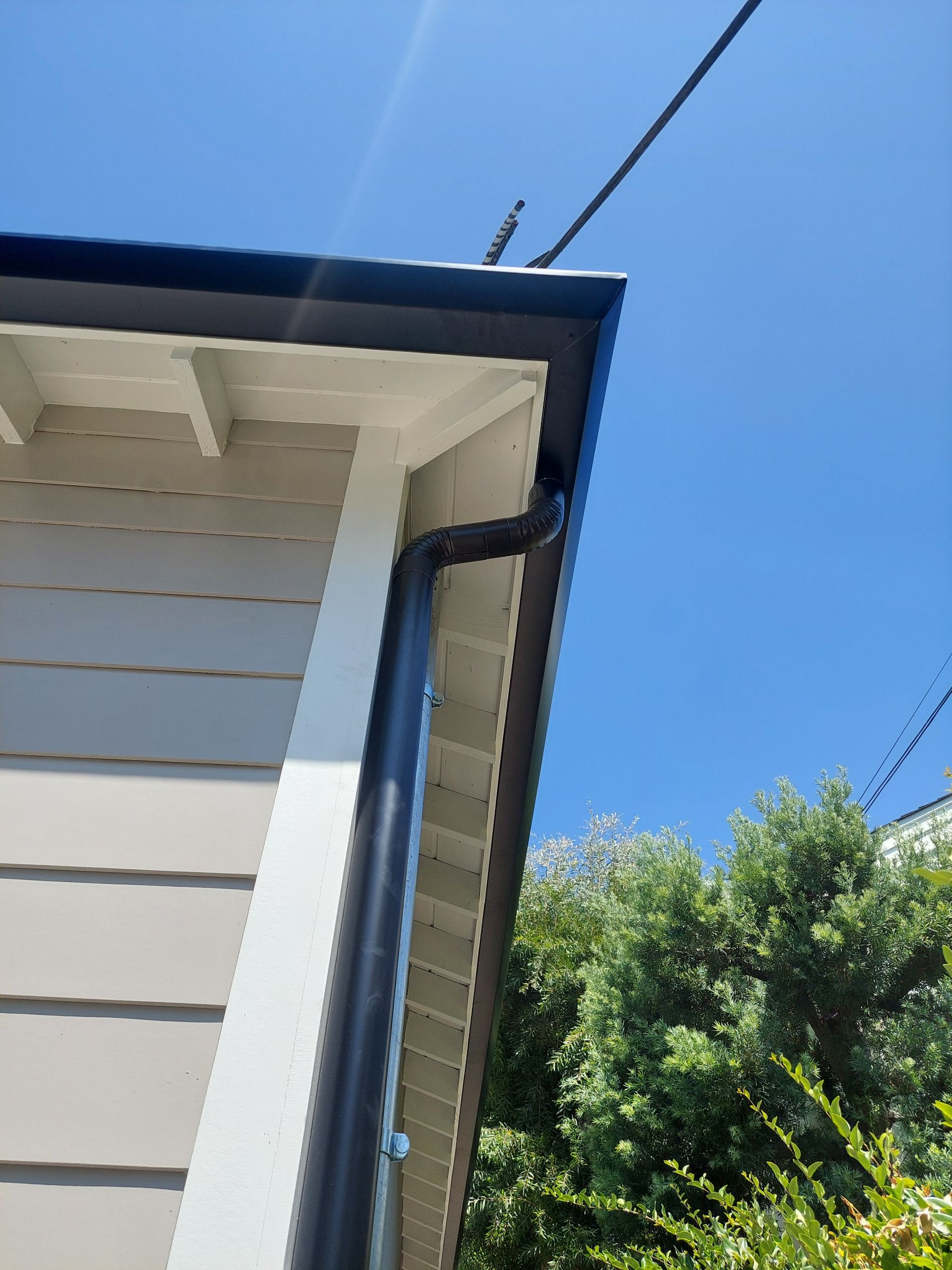 Corner of a white house with blue trim, downspout, and overhead power lines against a clear blue sky