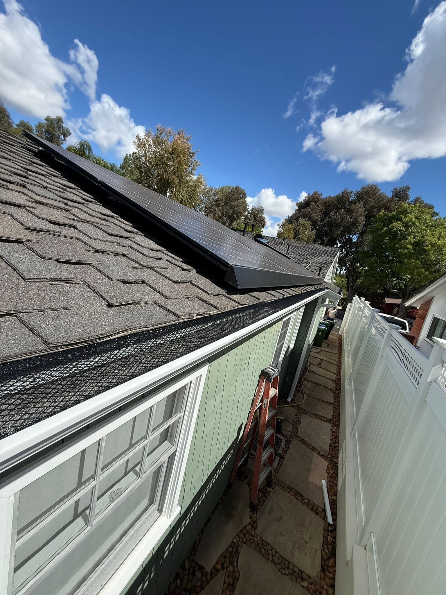 Narrow side yard beside a house with roof shingles, white fence, and blue sky with clouds