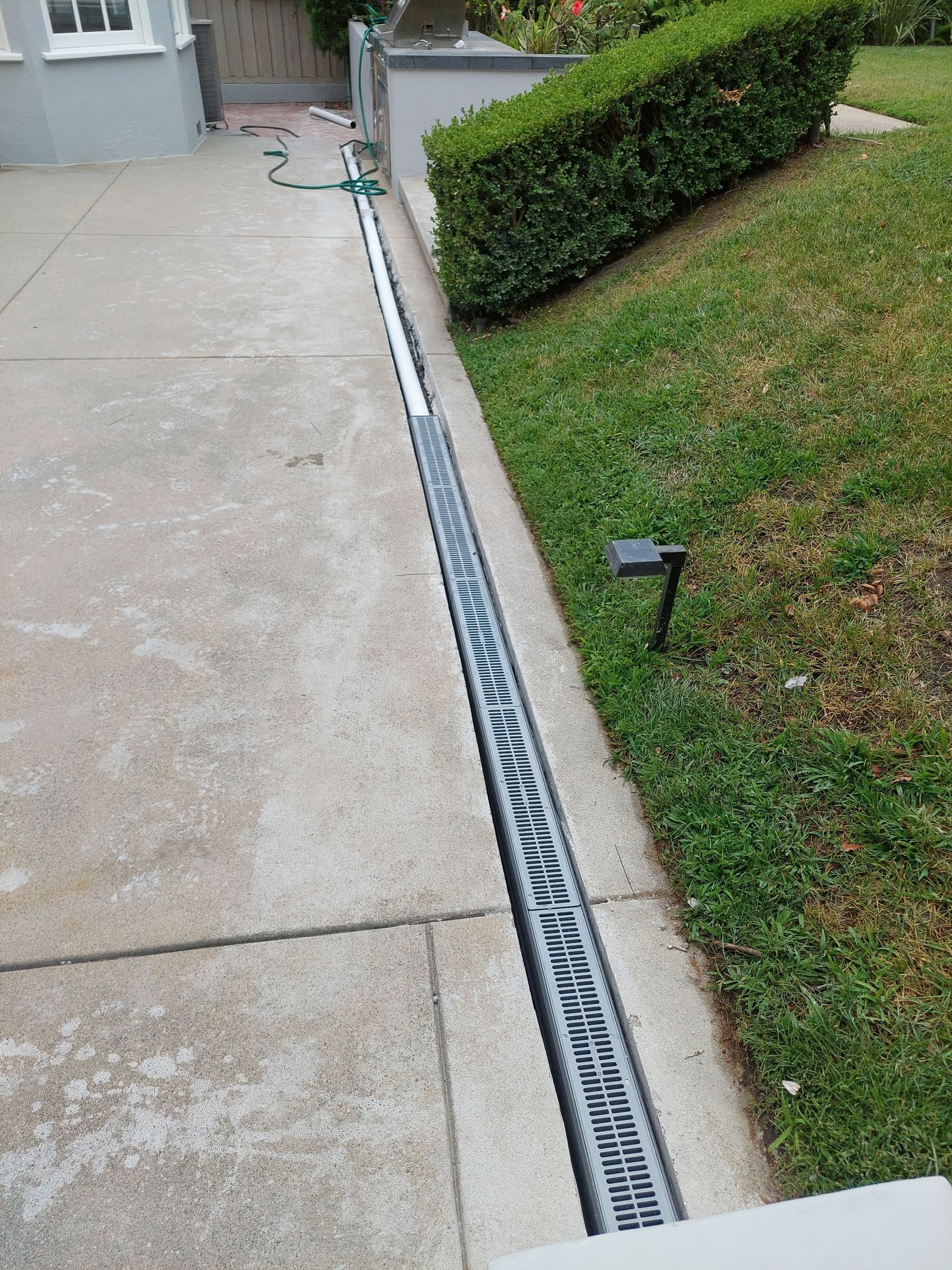 Concrete driveway beside grassy yard with a long metal drainage grate along the curb.